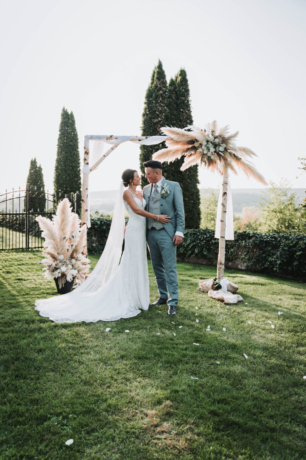 Braut und Bräutigam stehen verliebt unter einem mit Pampasgras geschmückten Traubogen im Garten bei Sonnenuntergang – romantische Outdoor-Hochzeit in Coburg.